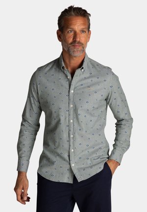 Man met baard, gekleed in een lichtgrijs overhemd met een blauw ruitpatroon en een donkere broek, staand met één hand in de zak.