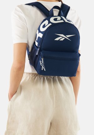 Person wearing navy blue Reebok backpack with white logo and lettering, paired with white short-sleeve shirt and beige shorts.