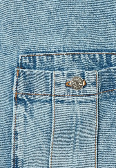 Tissu en denim bleu clair avec une texture visible. Comprend une poche avant, des coutures orange et un bouton argenté avec un détail de marque.