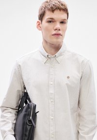 Πουκάμισο μακρυμάνικο με κουμπιά, σε ανοιχτό μπεζ και λευκές ρίγες, συνδυασμένο με μαύρη τσάντα με υφή, που διαθέτει φερμουάρ και χερούλι.