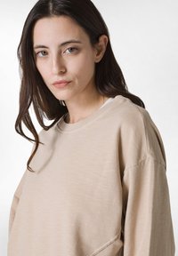 Sudadera de manga larga beige con cuello redondo y corte relajado. Suave al tacto, con costuras mínimas y hombros caídos que realzan la comodidad.