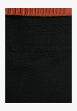 Tessuto nero con un motivo testurizzato, caratterizzato da una fascia in rib chevron arancione e cuciture verde scuro lungo la cucitura.