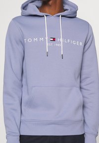 Felpa leggera viola chiaro realizzata in misto cotone, con tasca frontale, cappuccio con lacci e logo bianco "TOMMY HILFIGER" con una scatola di colore accentuato.
