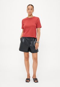 Rode katoenen T-shirt met korte mouwen, met knopenaccenten op de schouder; gecombineerd met zwarte faux leren shorts met hoge taille.