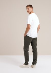 Witte t-shirt met korte mouwen, olijfgroene broek en beige sneakers; staande met handen in de zakken, iets naar achteren gekeerd.