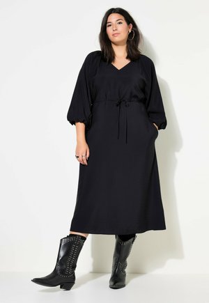 Robe maxi noire avec un décolleté en V, taille cintrée et manches bouffantes. Portée avec des bottes de cow-boy noires cloutées. Tissu lisse avec poches.