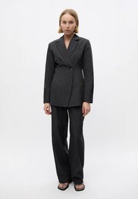 Mulher em pé vestindo um fato preto risca de giz com um blazer de botão único e calças largas, acompanhado de sandálias pretas de bico quadrado.
