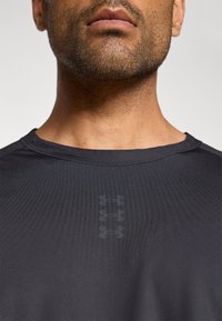 Μαύρο αθλητικό μπλουζάκι με στρογγυλή λαιμόκοψη, λεία υφή και διακριτικό λογότυπο σ ε σκούρο γκρι στο στήθος. Κοντά μανίκια.