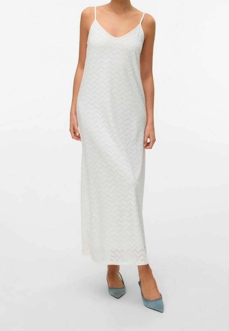 Femme portant une robe maxi blanche sans manches avec un motif œillet en chevron et des escarpins bleus à bouts pointus et talons bas, debout devant un fond blanc.