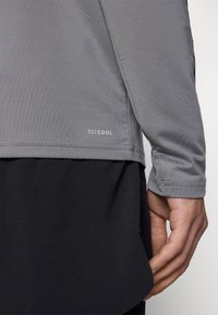Graues Sportoberteil mit strukturierter Oberfläche, versehen mit einem Logo mit der Aufschrift "BEECOOL", kombiniert mit einer schwarzen Schicht darunter.