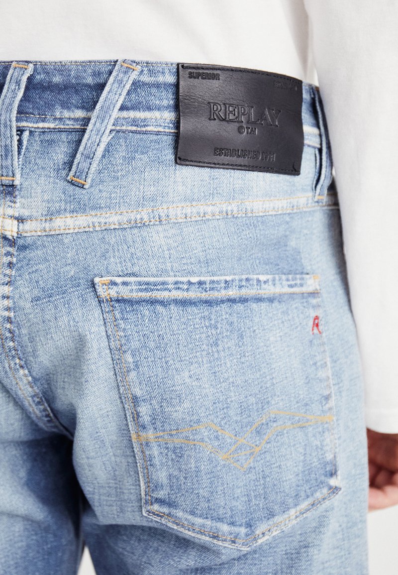 Hellblaue Jeans aus Denim mit einem Lederpatch, auf dem "REPLAY" geprägt ist. Auffällige Details sind der Kontraststich und eine Tasche mit Zickzackmuster.