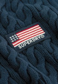 Temno modra pletena jopica z vrtnico in nalepko z ameriško zastavo ter napisom "SUPERDRY" v beli barvi.