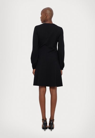 Schwarzes langärmliges Kleid mit tailliertem Oberteil und schwingendem Rock. Verfügt über einen hohen Ausschnitt und dezente Nahtdetails, kombiniert mit schwarzen High Heels.