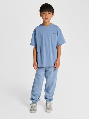 Junge mit glatten schwarzen Haaren, der ein blaues übergroßes T-Shirt, passende blaue Jogginghosen und silberne Turnschuhe trägt, steht vor weißem Hintergrund.