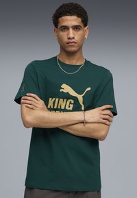 Ung mand med kort krøllet hår iført mørkegrøn Puma t-shirt med guld logo og tekst, arme krydset, sølv kædehalskæde og armbånd på grå baggrund.