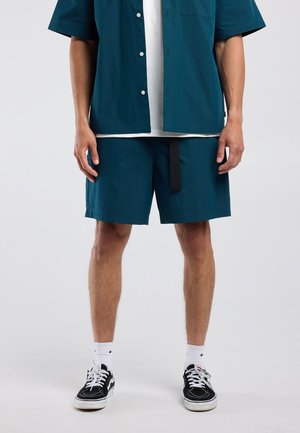 Mann trägt türkisfarbene Shorts und offen dazu passendes Hemd über weißem T-Shirt, weiße Socken und schwarze Sneakers, steht vor einfarbigem Hintergrund.