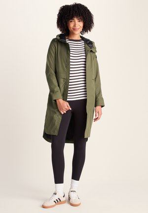 Young woman with curly hair wearing an olive green long coat, black and white striped shirt, black leggings, white socks, and white sneakers.