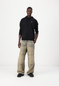 Čierna mikina s kapucňou a malým logom, kombinovaná so svetlými béžovými voľnými džínsami s viditeľnými švami a tónovými záplatami. Čierne tenisky.