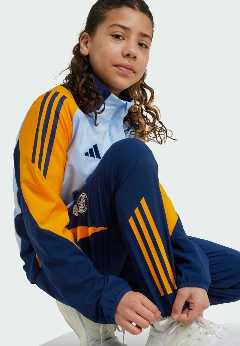 Track suit with blue and orange colors. Features three stripes on sleeves and legs, and logo on pants. Smooth texture and athletic fit.