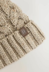 Beige strikket beanie med et struktureret, tykt kabelmønster og ribbede manchetter. Har et brunt læderakcent med et logo.