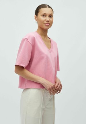 Krótki rękaw, top z dekoltem w serek wykonany z miękkiej, różowej tkaniny o subtelnej fakturze; w zestawie z jasnymi, luźnymi spodniami.