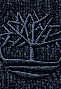 Il tessuto lavorato a maglia blu navy presenta un logo ricamato di un albero con rami intrecciati e una base curvata, evidenziando dettagli testurizzati.