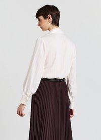 Blusa bianca a righe con polsini bottonati, abbinata a una lunga gonna plissettata marrone e una cintura nera in vita. Tessuto dall'aspetto liscio.