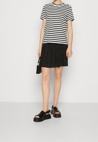 Person trägt ein kurzärmliges Oberteil mit schwarzen und weißen Streifen, einen schwarzen Faltenrock, schwarze Plateausandalen und hält eine schwarze Handtasche an der Seite.