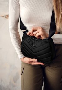 Černá prošívaná kabelka přes rameno s chevronovým vzorem, zipovým uzávěrem a popruhem. Materiál má hladkou texturu a elegantní povrch.