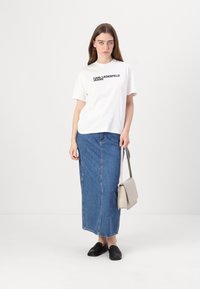 Biely tričko s čiernym textom "KARL LAGERFELD JEANS", skombinované s dlhou modrou denimovou sukňou, čiernymi mokasínami a svetlo šedou kabelkou cez plece.