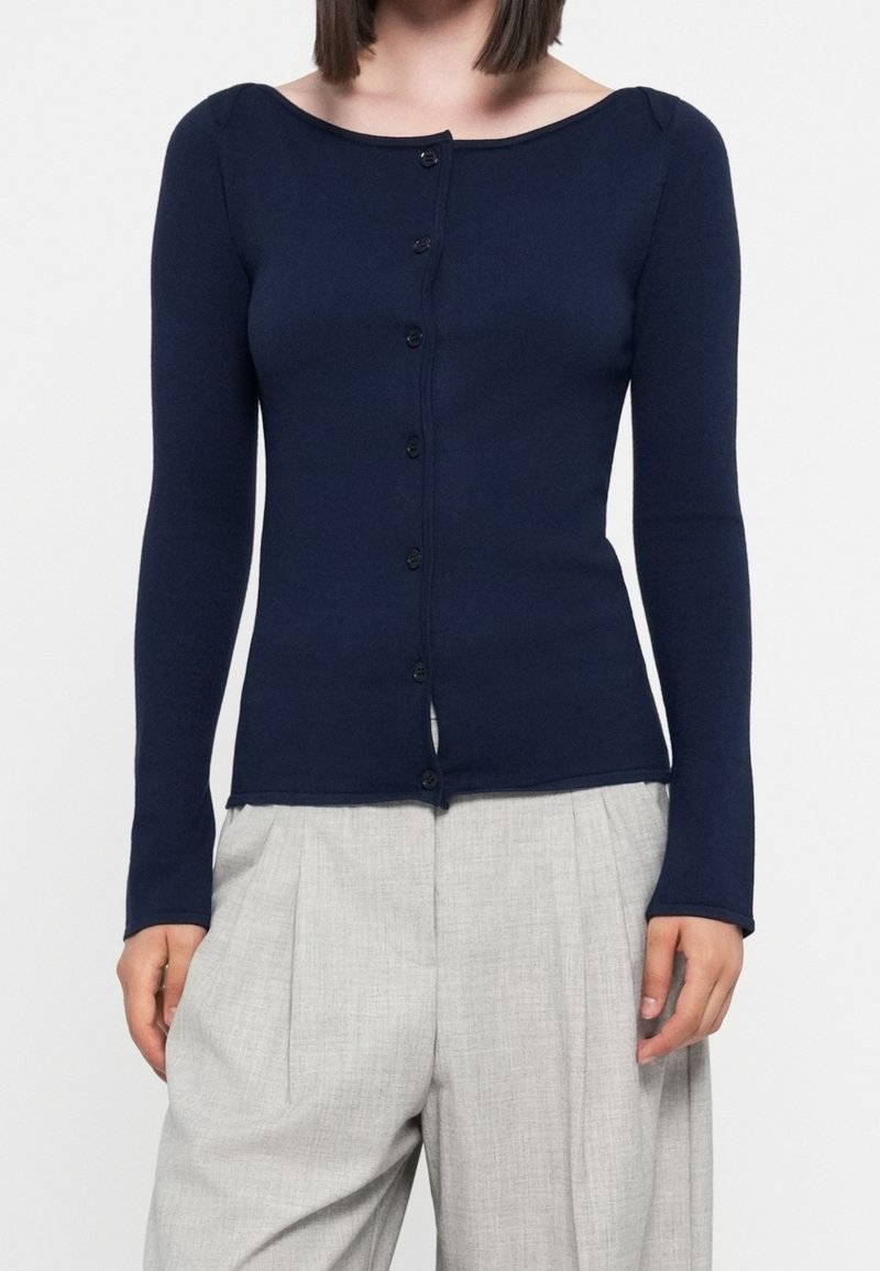 Femme portant un cardigan ajusté bleu marine à manches longues boutonné, associé à un pantalon plissé taille haute gris clair.