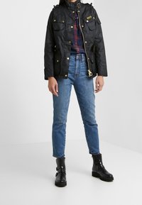Schwarze, wasserdichte Jacke mit goldenen Beschlägen, mehreren Taschen und Schulterträgern. Getragen über einem karierten Hemd und blauen Jeans mit schwarzen Knöchelstiefeln.