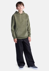 Oliwkowy, sznurowany hoodie z przednią kieszenią i ściągaczami, w połączeniu z luźnymi czarnymi spodniami i szarymi butami wsuwanymi.