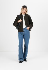 Ung kvinne står oppreist, iført en svart jakke, hvit T-skjorte, blå jeans, svarte sko, og bærer en svart skulderveske.