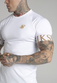 White short-sleeve t-shirt with a gold embroidered logo on the chest and a gold-accented band on the arm, fitted design, smooth texture.