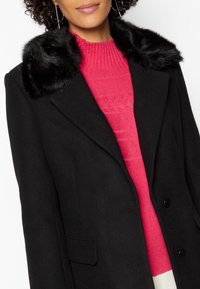 Abrigo negro con un suave cuello de piel sintética, diseño de doble botonadura, combinado con un suéter de textura rosa debajo.
