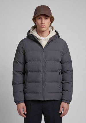 Jeune homme portant une casquette marron, un sweat à capuche beige et une veste matelassée gris foncé zippée, debout, bras détendus, sur un fond uni.