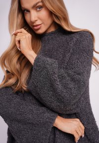 Szary, teksturowany sweter dzianinowy z długimi, szerokimi rękawami i wysokim dekoltem. Tkanina wydaje się miękka i przytulna. Brak widocznych wzorów lub ozdób.