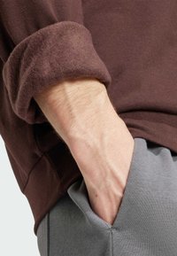 Brun skjorta med långa ärmar och uppvikta ärmslut, kombinerad med grå byxor. Tyget verkar mjukt och strukturerat med en ledig passform runt midjan.