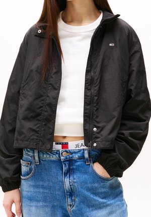Kvinde iført en sort lynlåstrøje over en hvid cropped top, blå jeans, med den ene hånd i lommen, der viser et mærke på linningen.