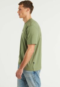 Žalia trumpa rankovė T marškiniai, pagaminti iš minkštos medžiagos, su apvali iškirpte ir logotipo printu priekyje. Suderinti su šviesiai mėlynomis džinsomis.