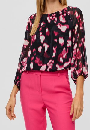 Femme portant un chemisier noir transparent à manches longues avec un imprimé abstrait rose et blanc, rentré dans un pantalon ajusté rose vif.