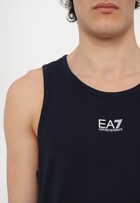 Tamsiai mėlynas medvilninis topas be rankovių su apvaliu iškirpimu, su baltu "EA7 Emporio Armani" logotipu. Sklandus paviršius, be rankovių dizainas.