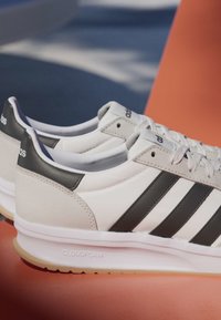 Białe trampki Adidas z czarnymi paskami i podeszwami "Cloudfoam" umieszczone na pomarańczowej powierzchni z rozmytym niebieskim tłem.