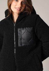 Veste en polaire noire avec une poche zippée au texture matelassée. Elle comprend une capuche et une fermeture éclair, ainsi que des poches latérales.