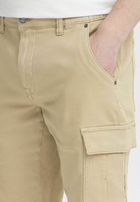 Pantalones cargo beige con un bolsillo lateral, cierre de botón y trabillas para cinturón. Hechos de algodón con una textura suave y un corte relajado.