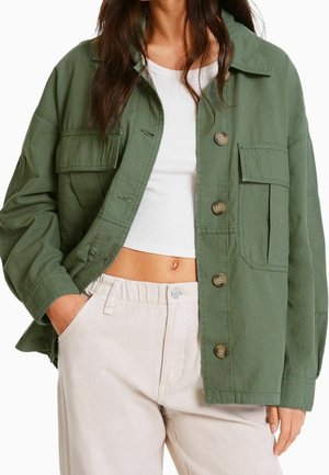 Femme portant une veste boutonnée oversize vert olive par-dessus un débardeur court côtelé blanc et un pantalon beige clair taille haute.