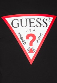 Zwarte T-shirt met een groot wit driehoekig logo met rode accenten en de tekst "GUESS USA WASHED JEANS" en een rode vraagteken.