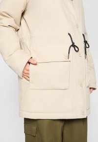 Veste matelassée beige avec une coupe décontractée, dotée d'une fermeture à boutons sur le devant, de grandes poches et d'un cordon de serrage ajustable à la taille.