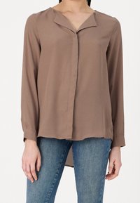 Brun langærmet bluse med v-udskæring, lavet af blødt stof. Har en knaplukning foran og en let buet kant, båret med blå jeans.